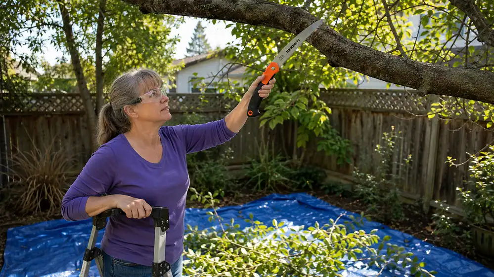 A hand gripping a folded pruning saw partway up a ladder, cutting through a thick branch